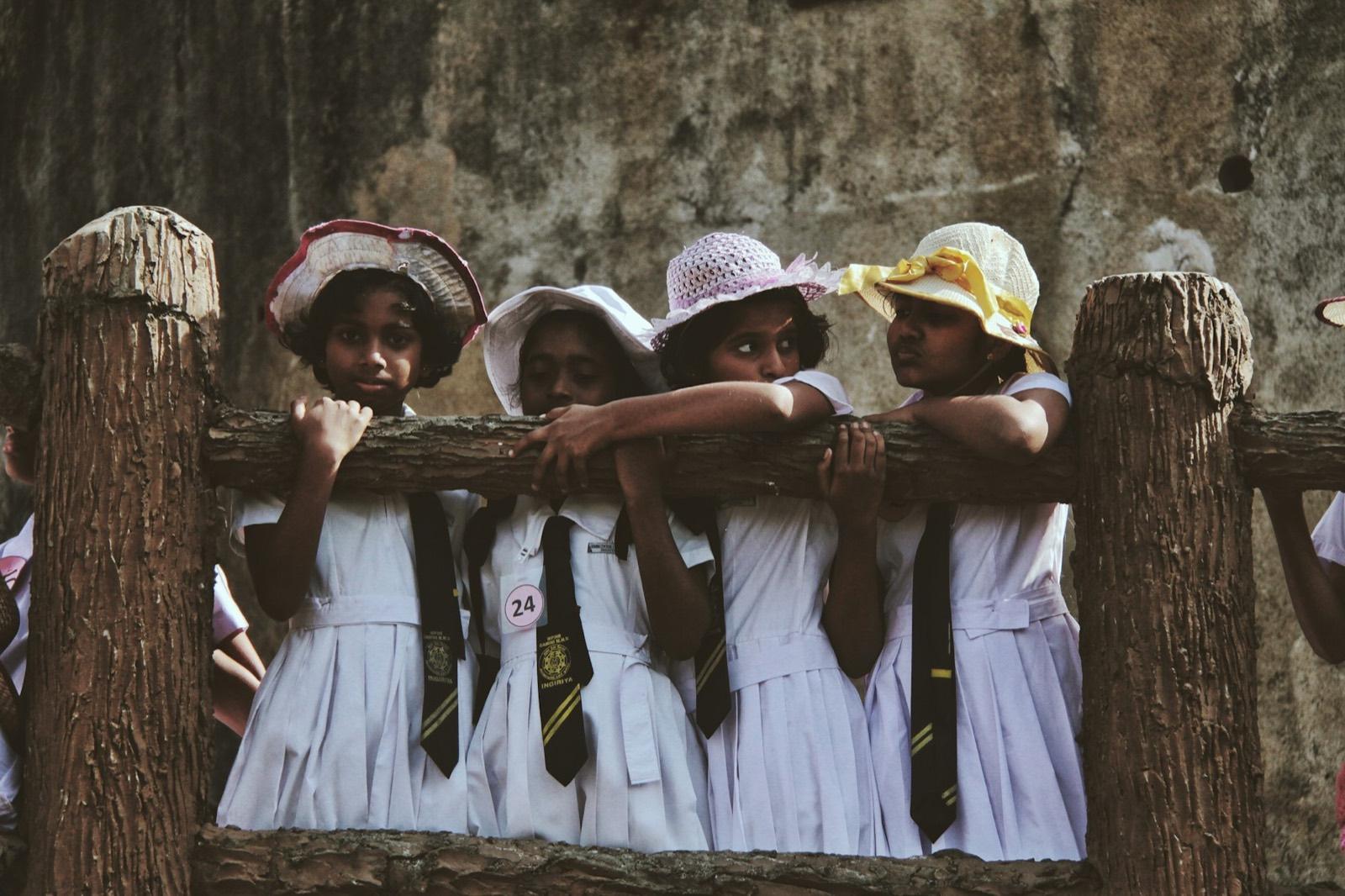 Schoolgirls in white uniforms and ties leaning on a wooden fence, Sabaragamuwa Province